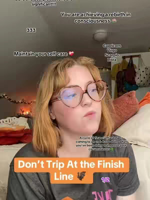 A selfie-style video cover image featuring a young woman with reddish-blonde hair and heart-shaped glasses. She is positioned in front of a bed with pillows and a patterned bedspread. Above her, there are text overlays with messages like "You are achieving a rebirth in consciousness" and "Maintain your self care." Further text includes the zodiac signs Capricorn, Virgo, Scorpio, and Libra. At the bottom, an orange banner reads "Don't Trip At the Finish Line" with a small bat symbol and the numbers "1/2."