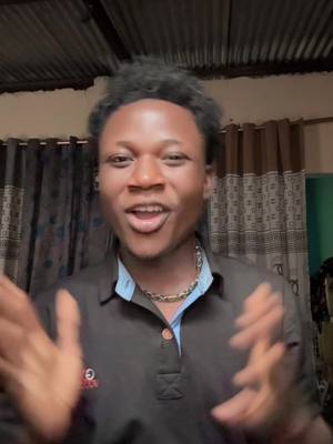 A young Black man with short, curly hair is centered in the frame mid-speech, wearing a dark polo shirt. The background, slightly out of focus, reveals a corrugated metal ceiling and patterned curtains. He gestures with both hands wide, conveying enthusiasm or emphasis.