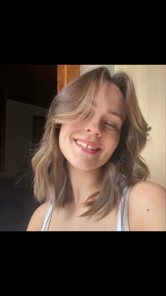 A close-up shot of a young woman with shoulder-length brown hair. Her eyes are closed, and she is smiling with her mouth open, revealing her teeth. She is wearing a white tank top. The background features a window and a wall with a light yellow color.