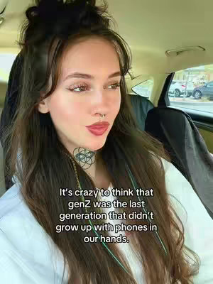 A young woman with long, dark hair, a nose ring, and a black head covering is seated in a car. She is wearing a white shirt and has a necklace with an intricate design.
