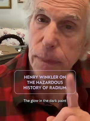 Image shows Henry Winkler in a close-up shot, partially visible. He's wearing a red and black plaid shirt, looking towards the camera with a serious expression. A text overlay in a translucent rectangle reads 'Henry Winkler on the Hazardous History of Radium' and below it 'The glow in the dark paint'. The background is slightly blurred, showing a room with a picture on the wall. The composition focuses directly on Winkler's face.