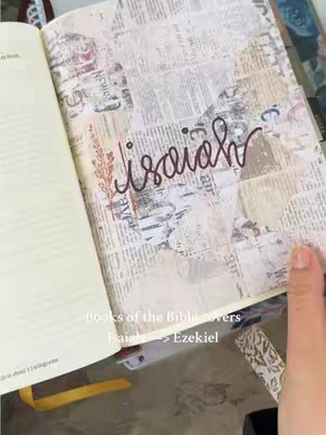 An open Bible with pages covered in a collage of newsprint, where the word "Isaiah" is handwritten, and adorned with a leaf-like design. A hand is visible in the corner, holding the page.