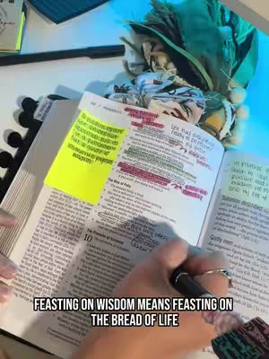 The image shows an open bible on a white surface. A hand with a ring is holding a black pen over the text, and areas of the text are highlighted in yellow and pink. A post-it note with text is attached to the bible page. The words 'FEASTING ON WISDOM MEANS FEASTING ON THE BREAD OF LIFE' are written at the bottom.