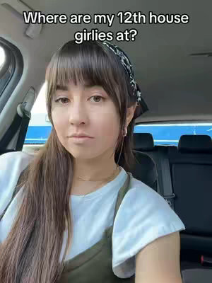 A woman with long brown hair and bangs is sitting in the driver's seat of a car and looking at the camera. The hook 'Where are my 12th house girlies at?' is overlaid on the image. She is wearing a white shirt and green dress and a black patterned headband. The car interior and the background are slightly blurred.