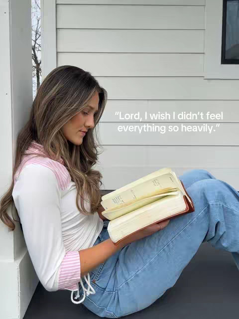 A young woman with long brown hair sits on a porch, leaning against a white wall. She is wearing a white shirt with pink striped accents and light blue jeans. She is holding an open book in her hands, with visible yellowed pages. The background shows a paneled white wall and some tree branches. Text overlay in white, includes a quote.