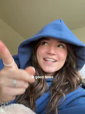 A young woman in a blue hoodie is pointing one finger towards the viewer, smiling. The text overlay says 'in good times'. The background is a light cream color.