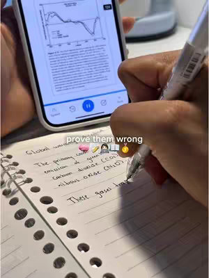 A hand with dark skin writing on a notepad. In the background is a smartphone displaying content. The notepad contains handwritten notes, along with the overlaid text "prove them wrong" and some related emojis.