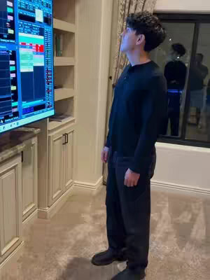 A man stands in front of a large screen displaying a trading platform, the background shows a home interior, featuring a wooden cabinet and a window with curtains.