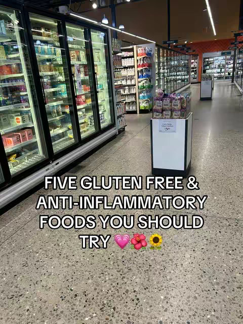 An interior shot of a brightly lit grocery store aisle. The focus is on the polished concrete floor with shelves of products on both sides and a sign in the middle. The text overlay reads the video title, 'FIVE GLUTEN FREE & ANTI-INFLAMMATORY FOODS YOU SHOULD TRY' in a bold, white font. Colorful emoji icons of hearts, flowers and a sunflower, are placed at the end.