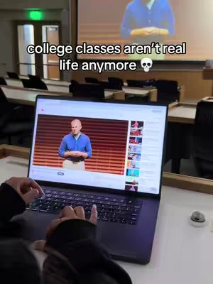 A person is sitting at a desk in a classroom, working on a laptop with a projected image on the wall behind the desk. The projection shows a man speaking in front of a simple background. The laptop is open, displaying what appears to be a video of a presentation. The hook text is white, in a sans-serif font, and appears on the upper left of the image. the classroom is slightly out of focus, highlighting the laptop and the text.