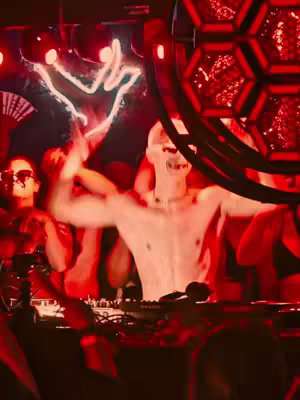 The image captures a dynamic scene at a nightclub or rave, bathed in an intense red light. A central figure, seemingly the focus of the scene, is a shirtless individual wearing a white, stylized mask covering their face. Their arms are raised, suggesting a high-energy moment. The background is filled with a crowd, partially visible, and a complex lighting rig enhances the atmosphere. The composition is dark, with the red light creating high contrast and highlighting elements of movement and excitement.