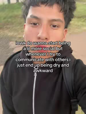 A selfie-style shot of a young man, framed from the chest up. He is looking at the camera with a neutral expression, standing outside. Text overlayed on the image reads: "lowk do wanna start being a lil more social but whenever I try to communicate with others i just end up being dry and awkward"
