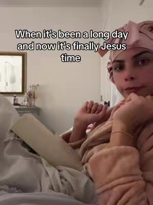 A woman wearing a pink head covering and a robe is lying in bed, holding a book. The background has a mirror and a small cabinet. The lighting appears soft and natural, and the overall feel is one of relaxation.