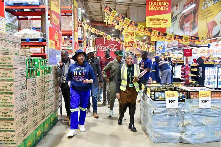 An interior shot of a brightly lit supermarket is filled with shoppers and staff, amidst high shelves stacked with products and promotional banners. A group of people, including a woman wearing a navy blue hoodie, blue pants with yellow accents, and a blue hat, walk towards the viewer. Others are wearing uniforms, some using electronic devices, appearing to be government officials or enforcement personnel.