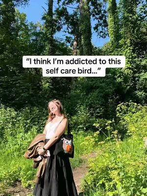 A woman stands on a path in a forest-like setting, with a white text box overlay above her head. The background consists of lush greenery and tall trees.
