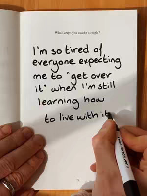A page from a notebook lying open on a wooden surface. The left side is blank, while the right side displays handwritten text in thick black marker. A hand holding a marker is visible in the lower right, about to continue writing. The text reads, 'What keeps you awake at night?' followed by, 'I'm so tired of everyone expecting me to "get over it" when I'm still learning how to live with it.' The overall composition implies introspection and personal reflection.