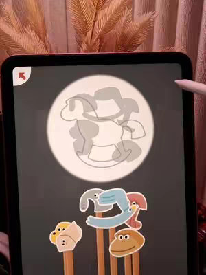 The image shows an iPad displaying a puzzle game. The game shows a silhouette of a rocking horse in a circle with a directional arrow. Various cartoonish animal heads on sticks are lined up below. The background has muted pink-toned elements and some dried flowers. The overall composition is neat, with clear focus on the game screen.