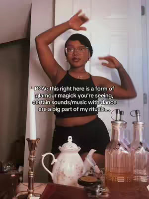 A woman with dark skin, wearing glasses and a black tank top, is dancing with her arms raised in a room. Various bottles and an ornate teapot are in the foreground.