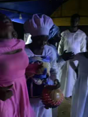 A group of people in white and pink robes are gathered indoors. A woman in a pink dress is in the foreground, and others in white clothing are dancing and clapping. There is a sense of celebration or religious ceremony. The lighting is dim, giving the scene a warm tone.