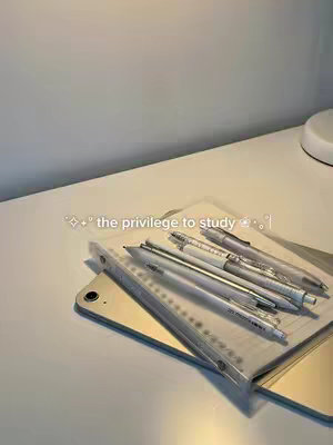 A well-lit, top-down shot showcases a white desk with a gold iPad and a notebook covered in pens. The pens are neatly arranged, with a mix of silver and transparent barrels. The composition is clean and focuses on study supplies.
