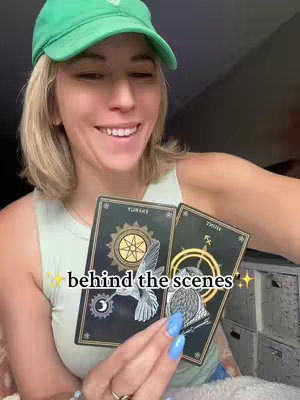 A woman in a green cap is holding tarot cards, smiling at the camera. Text overlayed on the image says 'behind the scenes'. The card on the left has the word 'ENERGY' and a hummingbird image on it. On the right is a card with the word 'HINT' with an owl on it. The scene has a neutral background.