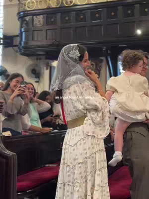 A woman in a white dress and veil is standing in a church, smiling and holding her hand near her face, with the text overlayed on the image: “an angel found her wings.” Other people can be seen in the background.