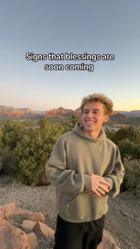 A smiling young man with curly hair wearing a green hoodie stands on a rocky outcrop with a scenic mountain view in the background, text overlayed on top.