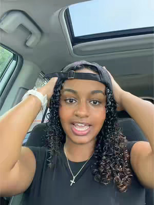 A selfie taken inside a car. The person is a young woman with dark curly hair, wearing a black cap, a black shirt, and a white watch. Her hands are on her head. The car has a sunroof and the background is blurry.