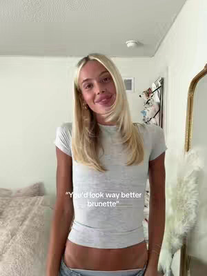 A woman with long, layered blonde hair stands in front of a light-colored wall and an ornate gold mirror. She is wearing a light gray t-shirt and jeans, facing the camera with a slight smile. Text overlayed gives a dialogue hint.