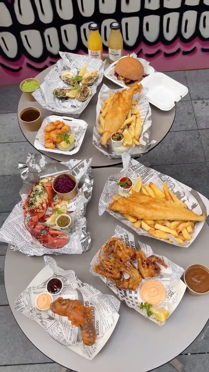 A top-down shot of a table filled with various dishes of food, including fish and chips, a lobster, oysters, and a burger, arranged on white paper with newspaper print. The table appears to be outdoors against a wall with a pink and white design. The overall composition is vibrant and appetizing.