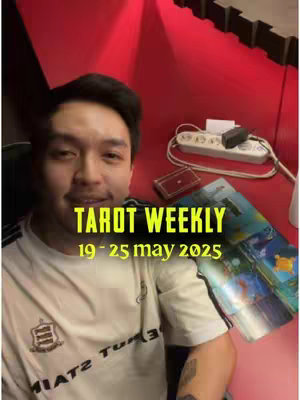 A man is smiling at the camera in a room with red walls. Text on the image says, "TAROT WEEKLY" and "19 - 25 may 2025". There's a deck of tarot cards, a power strip, and a laptop visible.