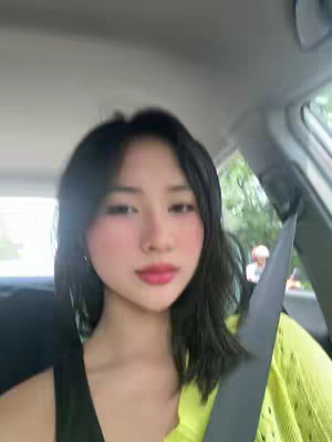 A selfie taken from inside a car, focusing on a young woman's face. She has long, dark hair and is wearing eye makeup with pink blush and red lipstick. A seatbelt crosses her body, and a portion of the car's interior and the outdoor scenery are visible in the background.