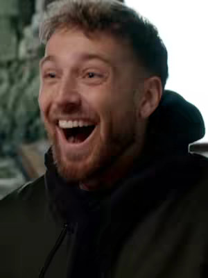 A close-up shot of a man with fair skin and light brown hair, his mouth wide open in a laugh, displaying his white teeth. He has a neatly trimmed, reddish-brown beard and is wearing a dark hoodie.
