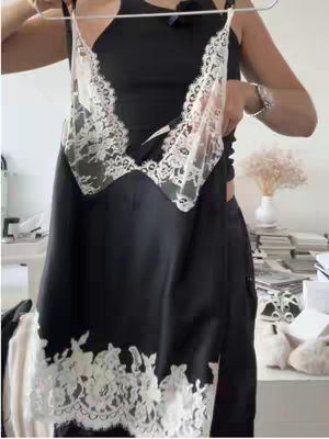 A person holds up a black slip dress with white lace detailing. The dress is hanging on a hanger. The background is a light-colored wall and a table with items on it.