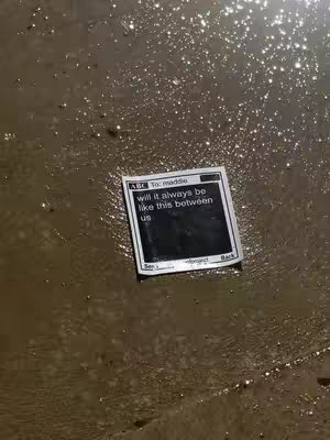 The cover image displays a close-up shot of a wet, reflective surface, likely water or a similar liquid. A rectangular paper is positioned slightly off-center, floating partially submerged. The paper suggests a mobile phone interface, featuring a text message dialogue bubble with the words; Will it always be like this between us?. The color palette is muted, with shades of brown and gray dominating the scene because the lighting is dim.