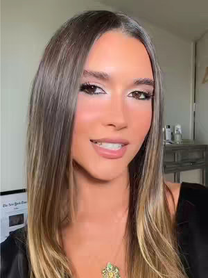 A close-up shot of a woman with long, highlighted hair and full makeup, including defined eyebrows, eye shadow, and lipstick. She is smiling slightly, and there's a necklace visible at the neckline of her black top. A background shows a room.