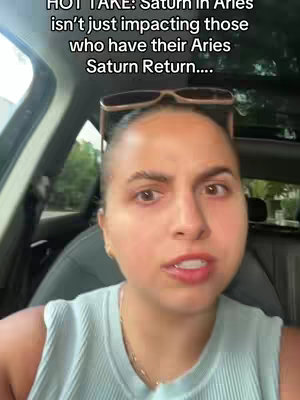 A woman with dark hair and sunglasses sits in a car, looking towards the camera and speaking. Text on the screen reads: HOT TAKE: Saturn In Aries isn't just impacting those who have their Aries Saturn Return....