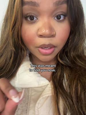 A selfie-style image of a woman looking directly at the camera. She has long, wavy brown hair with lighter highlights, and a neutral expression with a slight pursing of her lips. She's wearing a cream-colored shirt. Text overlay sits in the middle frame, with the words 'This was meant to find you now.' The background appears to be a neutral-colored wall.