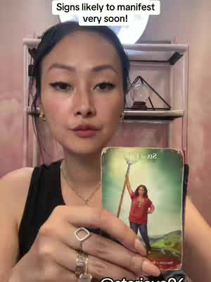 A woman with dark hair holds a tarot card, looking directly at the camera. Text at the top reads, 'Signs likely to manifest very soon!'. The woman is in a softly lit room with a shelf containing a decorative sphere and a small light. She wears a ring and a necklace.