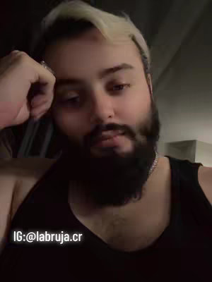 A medium shot of a man with lightened hair on top and a bushy, dark beard. He's wearing a black tank top and resting his head on his hand, with a slightly weary expression. The background is dim, but a glimpse of a room can be seen.