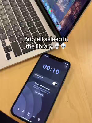 The cover image shows a smartphone on a wooden surface with a timer on its screen, next to a laptop with the screen open showing a program, all set in what appears to be a library setting. The text overlay reads Bro fell asleep in the library with two skull emojis.
