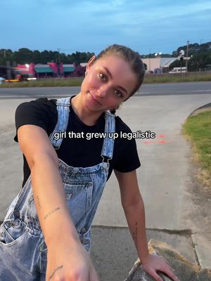 A young woman with brown hair and a black t-shirt under blue overalls leans toward the camera, resting on a curb near a road. The background shows a road, buildings, and twilight hues.