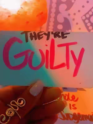 A hand holding a sign that says 'They're Guilty' in handwritten, pink letters. The background has a teal and pink ombre effect. The nails are painted white and the hand is wearing a silver ring.