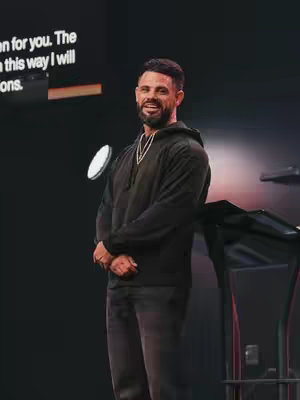 The image depicts a man in a black hoodie standing in front of what appears to be a dark stage or setting with a podium. He has short, dark hair and a beard, and he is smiling. There is text visible in the upper-left corner, partially obscured, and a circular light source.