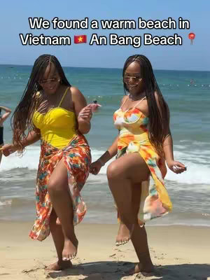 Two women in swimwear are on a beach, smiling and holding hands. They are standing on the sand with the ocean and sky in the background.