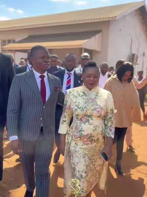 A group of people walking outdoors, including two main figures - a man in a pinstripe suit and a woman in a floral dress. Other individuals are visible in the background, some with suits and ties, suggesting a formal or professional setting. The lighting appears bright, and the overall composition gives a sense of movement.