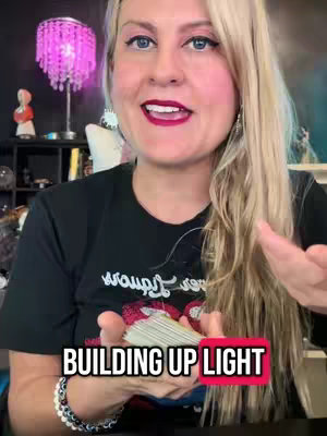 A woman with long blonde hair and fair skin, wearing a black t-shirt and a beaded bracelet, is holding a deck of cards in front of her. The background includes a pink, ornate lamp and bookshelves with various items. The text overlay reads 'BUILDING UP LIGHT'.