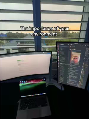 The image captures a workspace with a strong emphasis on environment. Two large computer monitors dominate the scene, with a laptop positioned below. The monitors display content, and the open laptop shows a colorful screen with an app. Blinds in the background partially obscure the outside view. Above the monitors, a text overlay reads 'The importance of your environment'.