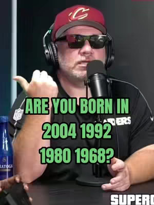 A man wearing sunglasses and a baseball cap sits in front of a microphone, with text overlayed on the image and appears to be talking. The background is dark.