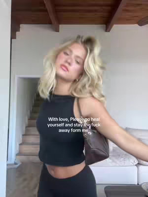 A woman with long, wavy blonde hair is captured in a medium shot, set within a bright, neutral-toned interior. She wears a black crop top and leggings, and is carrying a brown shoulder bag. The image has a bright and airy feel, enhanced by the lighting from the ceiling and the bright background.